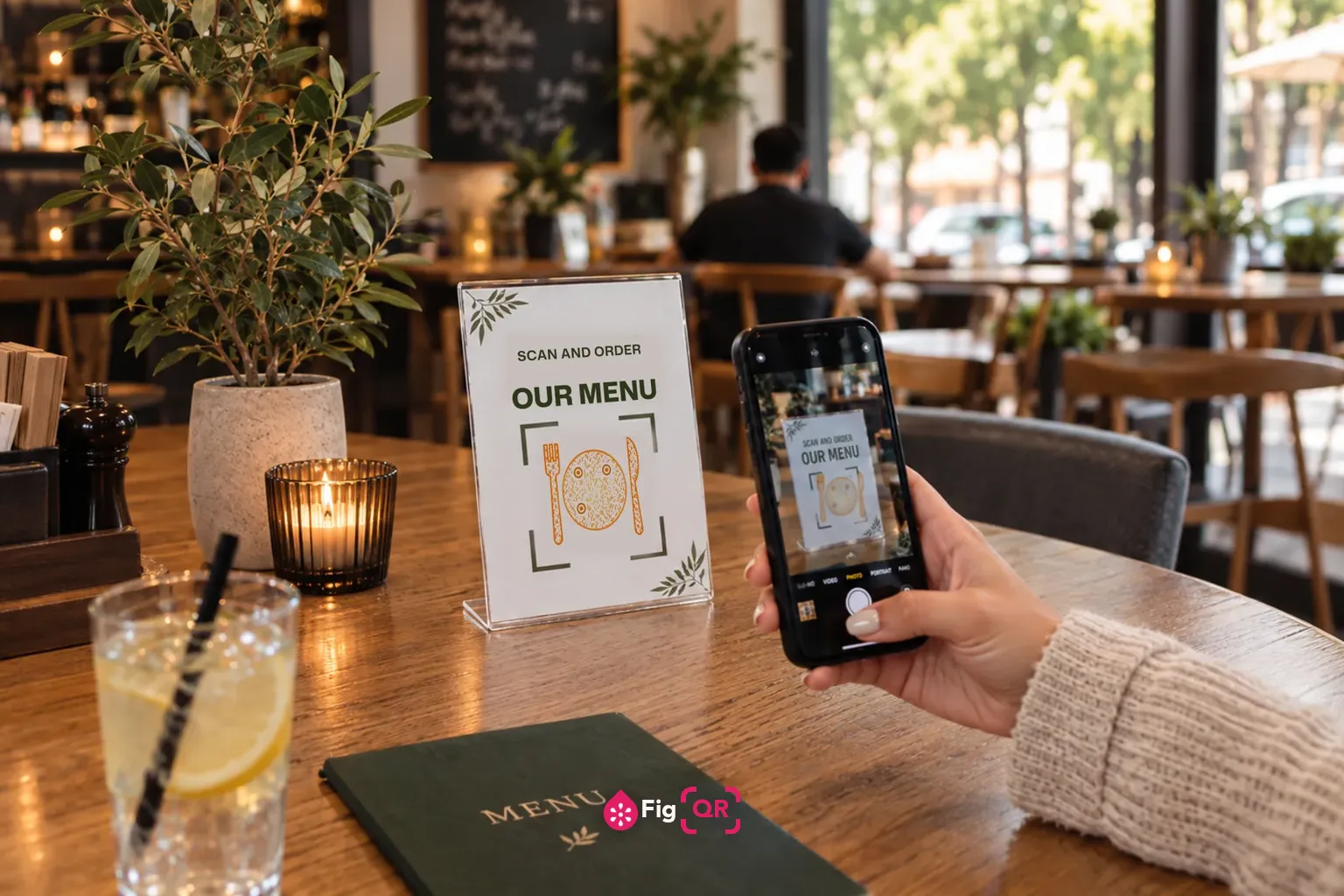 A menu card standing on a café table, printed with SCAN AND ORDER OUR MENU above a QR code drawn as a plate with a fork and knife, while a diner holds a phone up to scan it.